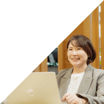 グレーのジャケットを着た女性がノートPCを操作しながら笑顔で話す様子