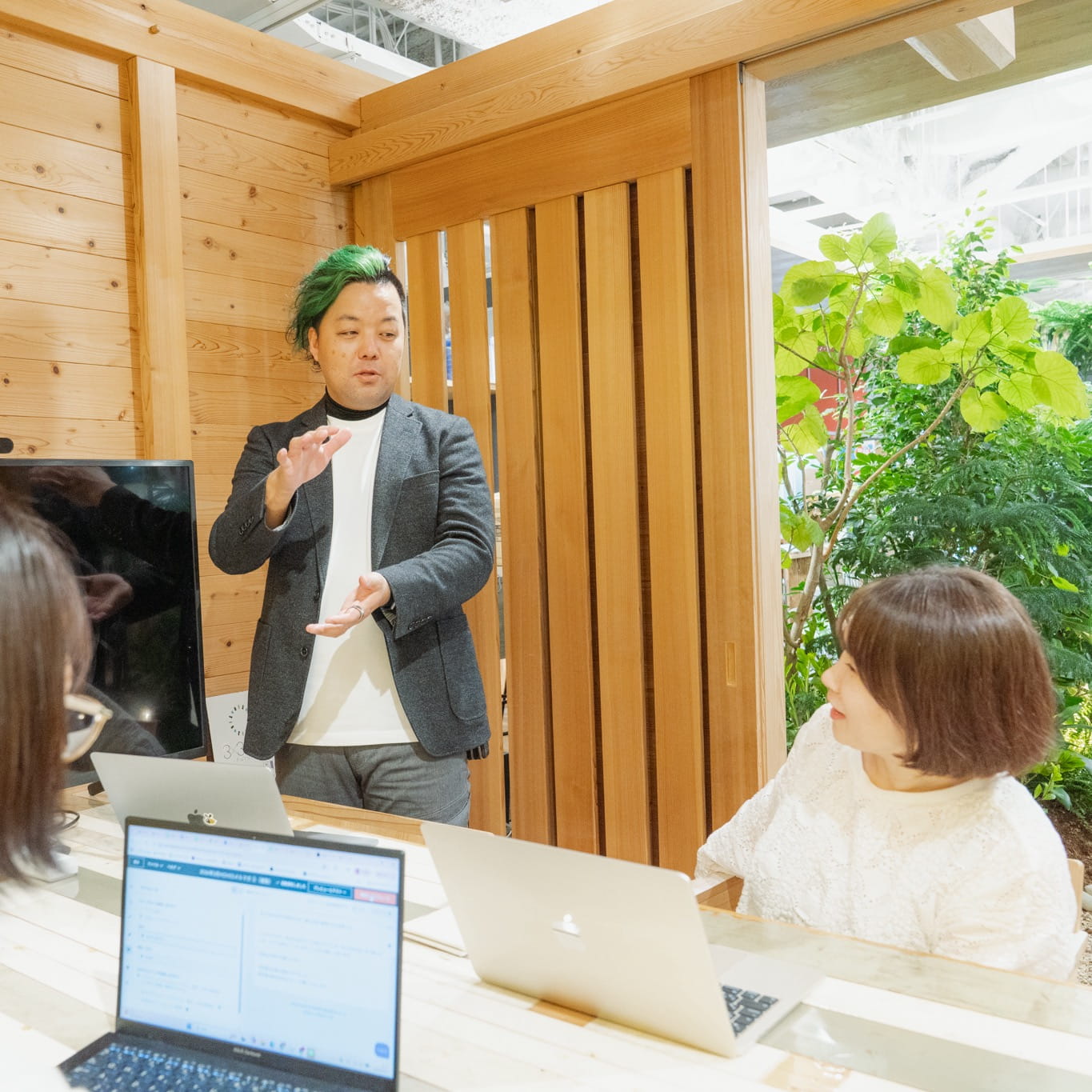 木製インテリアのオフィスで、男性が他の社員に向けて身振り手振りを交えて話をしている様子